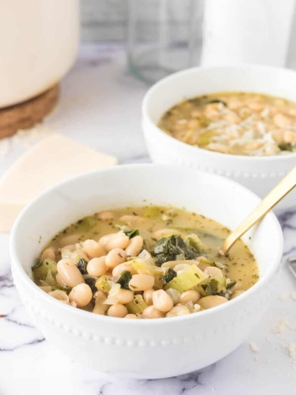 Two white bowls filled with white bean soup featuring greens and herbs. The soup is garnished with a sprinkle of cheese. A gold spoon rests in the foreground bowl. The setting includes a marble surface and a blurred background, highlighting the comforting dish.