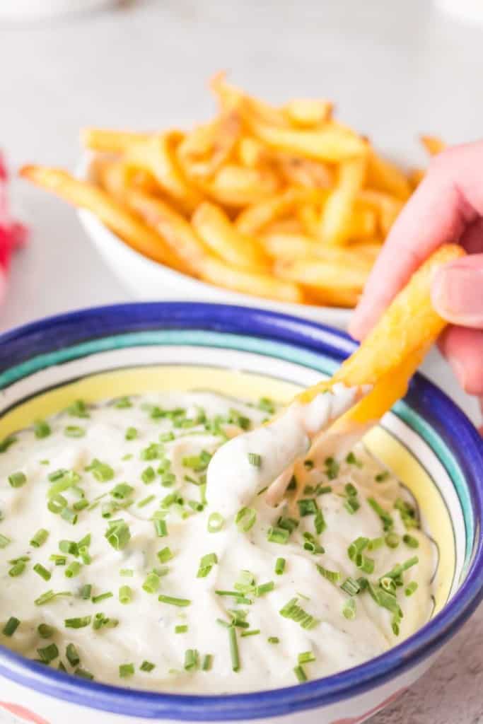POV dipping fries into a small bowl of aioli with chives on top