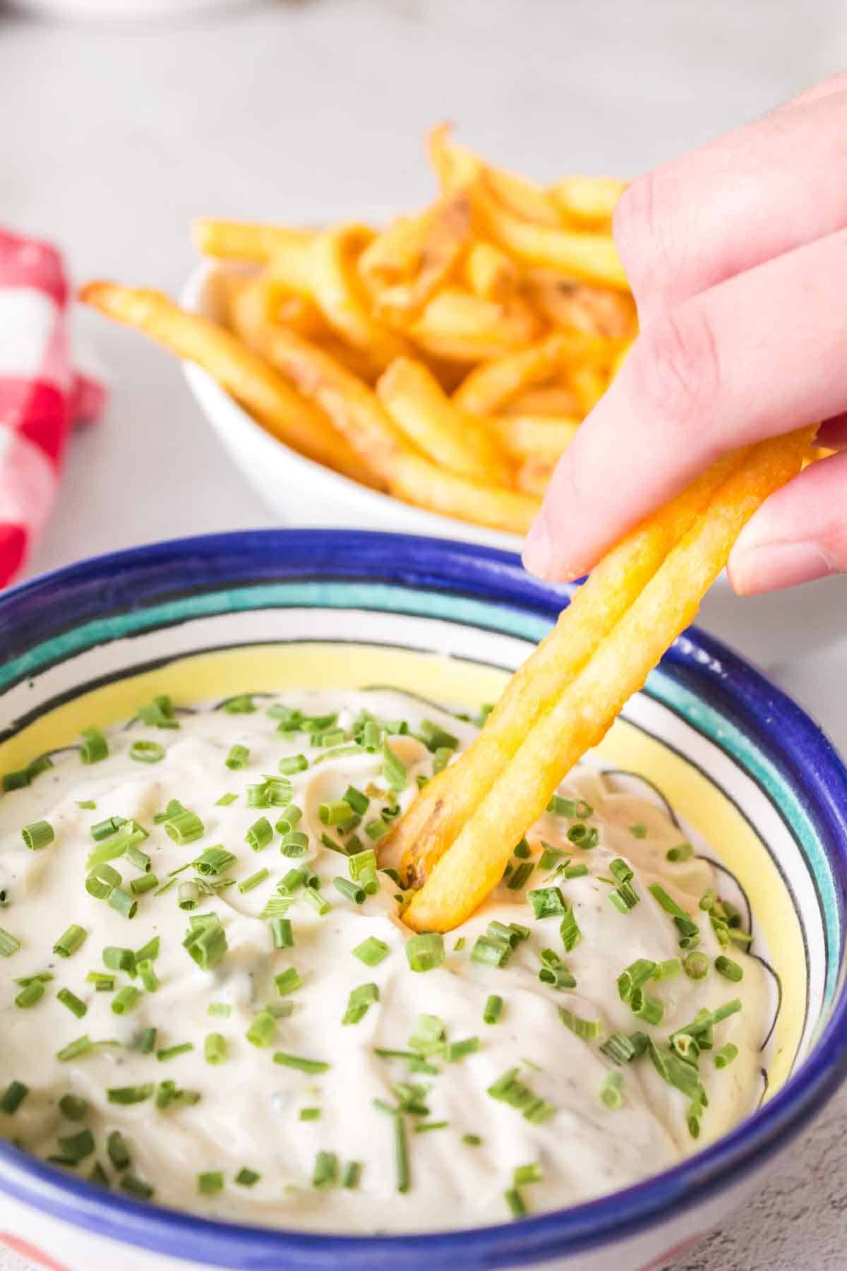 POV dipping fries into a small bowl of aioli with chives on top