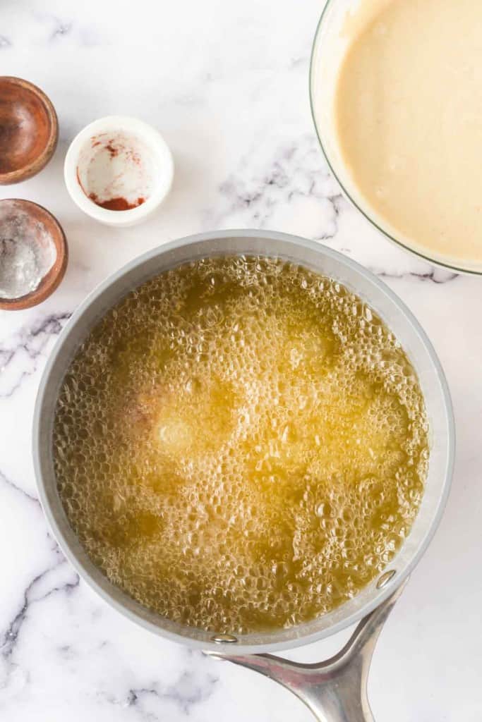 A saucepan filled with bubbling oil on a marble surface awaits the golden crisp of chicken and waffles. Surrounding it are small bowls containing spices and a large bowl with creamy batter, ready to craft this savory-sweet delight.