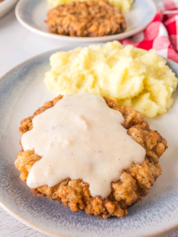 A plate of chicken fried steak, crafted from a timeless chicken fried steak recipe, is lavishly covered in white gravy and served with a generous portion of mashed potatoes. The dish is elegantly arranged on a light gray plate, with a red and white checkered cloth peeking from the background.