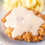 A plate featuring chicken fried steak smothered in creamy white gravy sits beside a generous serving of mashed potatoes. This classic dish is elegantly presented on a light-colored ceramic plate atop a pristine white surface.