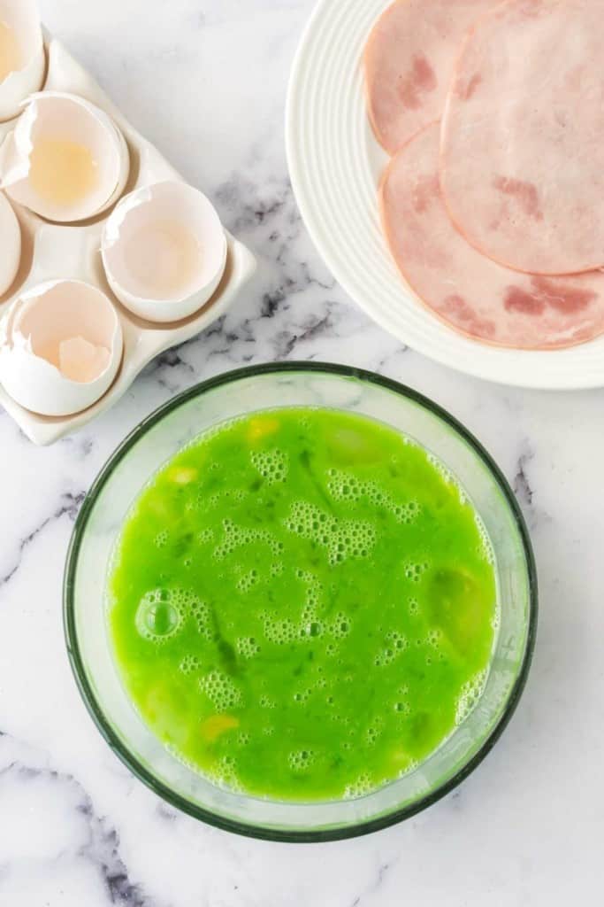 green colored raw eggs beaten in a mixing bowl with ham on the side