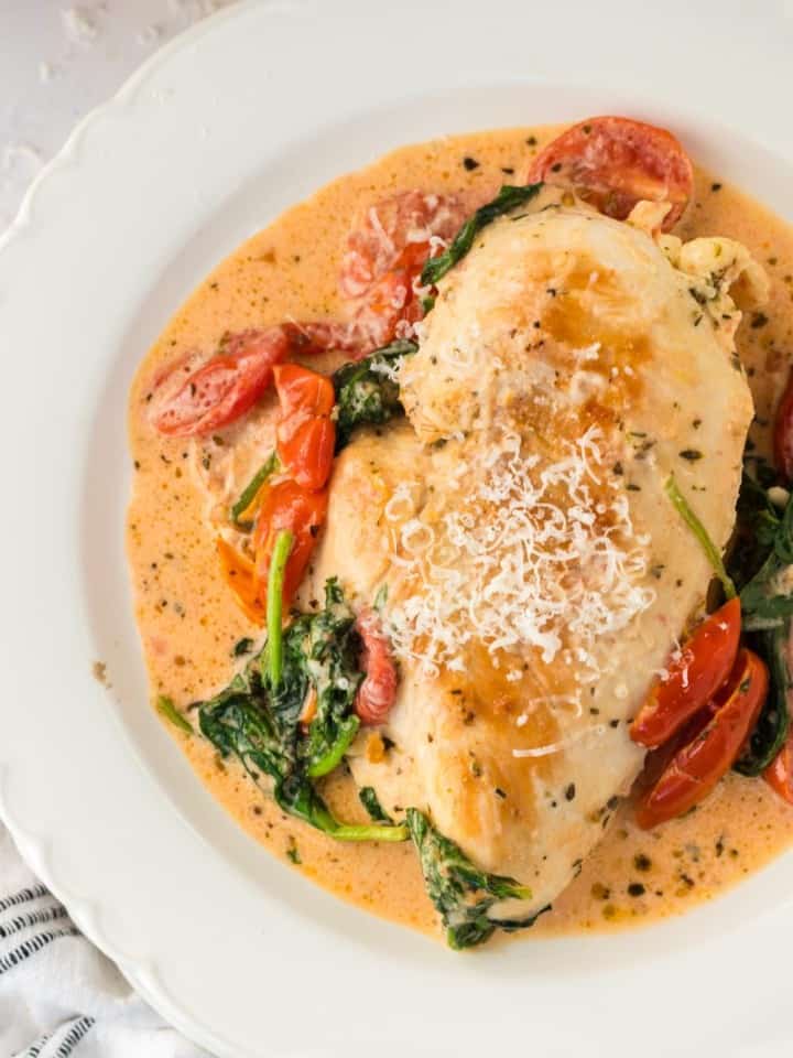 creamy tuscan chicken recipe with a whole breast in sauce in a white bowl garnished with tomatoes and greens