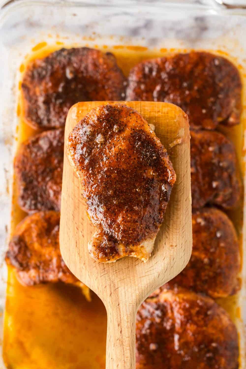 a wooden spatula over a clear baking dish with seasoned pork chops in a sauce