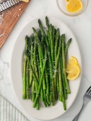 a white plate with sautéed asparagus with parmesan cheese and lemon on the side
