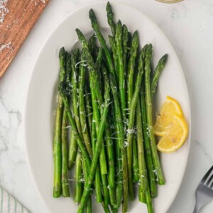 a white plate with sautéed asparagus with parmesan cheese and lemon on the side.