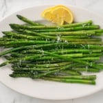 a white plate with sautéed asparagus with parmesan cheese and lemon on the side.