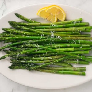 a white plate with sautéed asparagus with parmesan cheese and lemon on the side