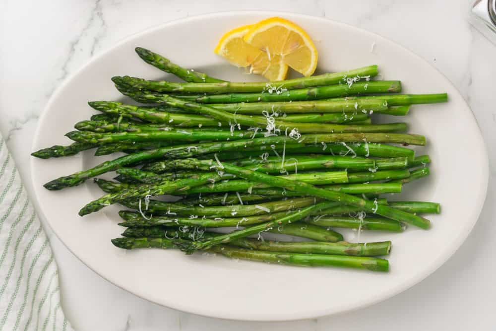 a white plate with sautéed asparagus with parmesan cheese and lemon on the side.