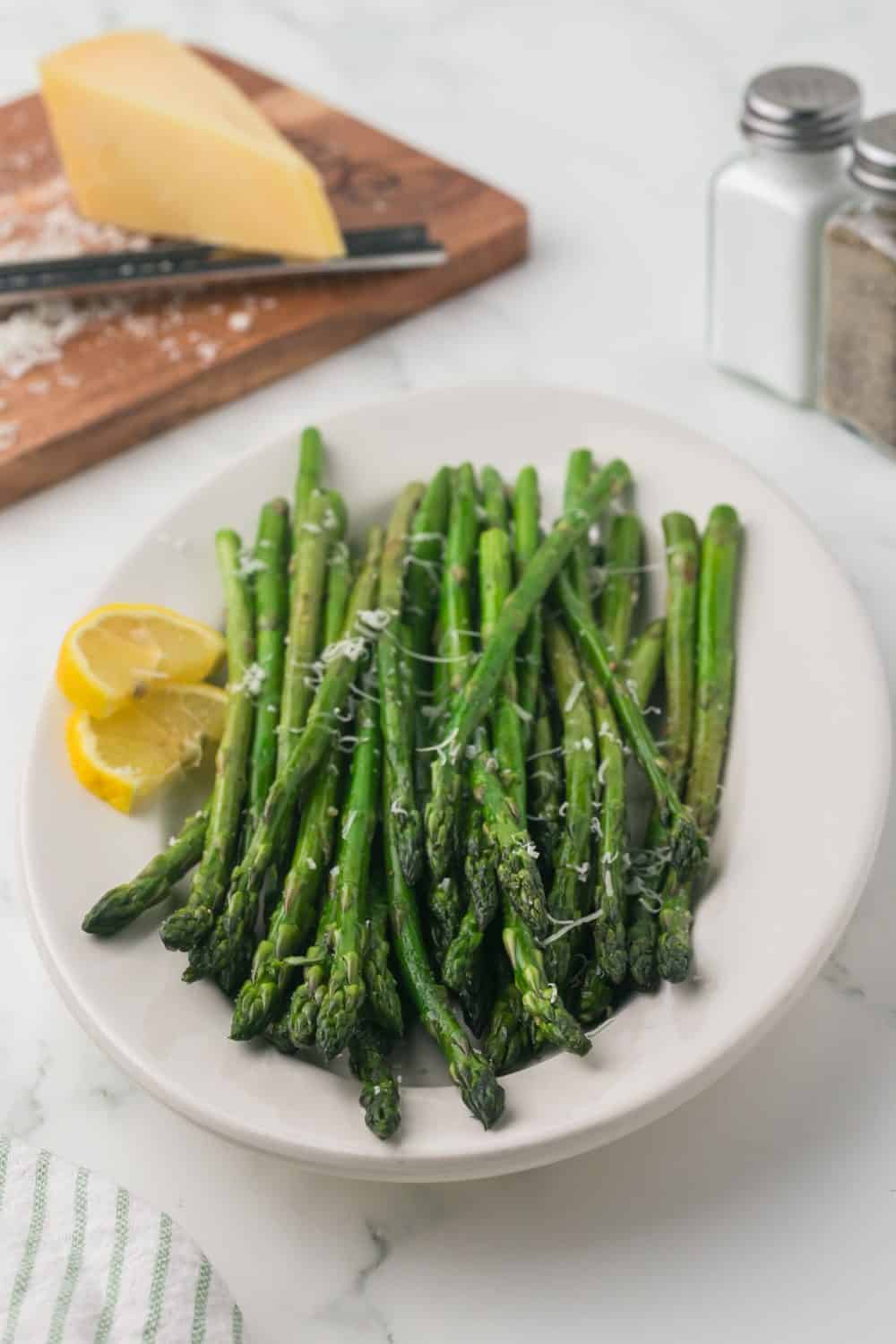 a white plate with sautéed asparagus with parmesan cheese and lemon on the side