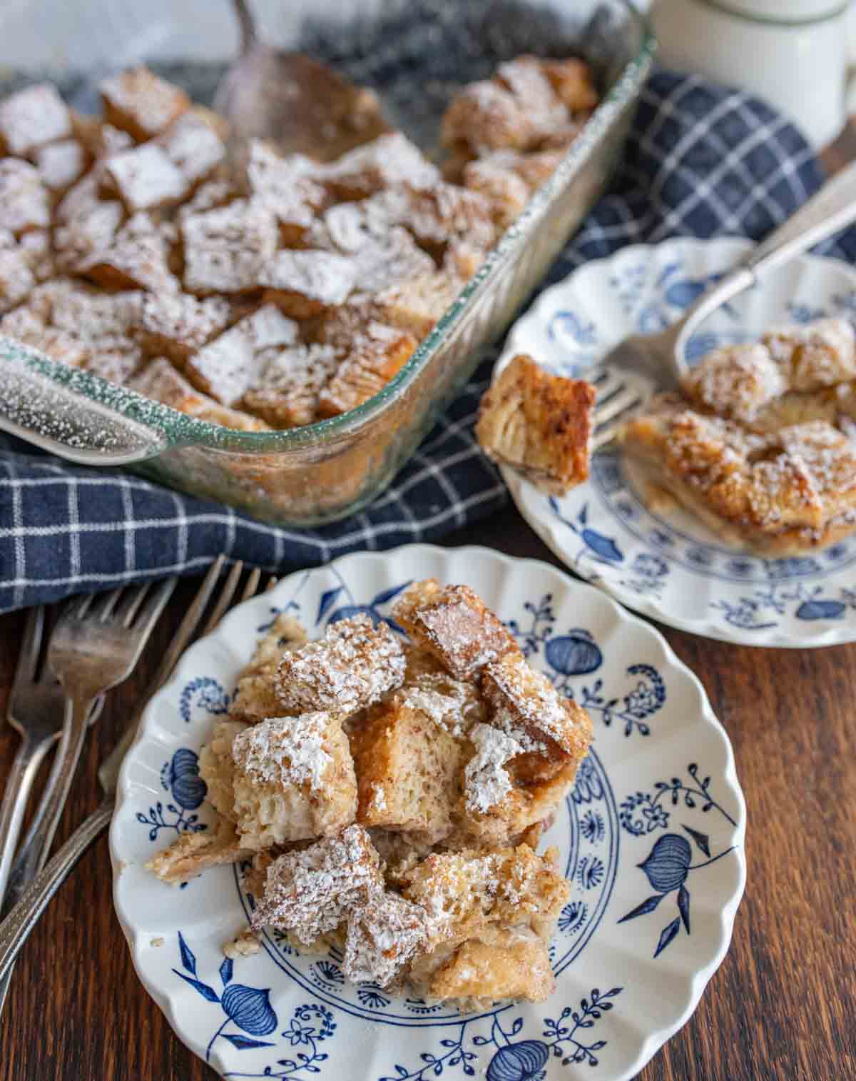 clear baking dish of squares of bread pudding with powdered sugar atop and served onto classic china dishes on the side