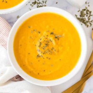 bright orange carrot soup in white serving bowls with herbs and salt and pepper to the side