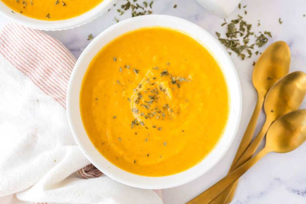 bright orange carrot soup in white serving bowls with herbs and salt and pepper to the side