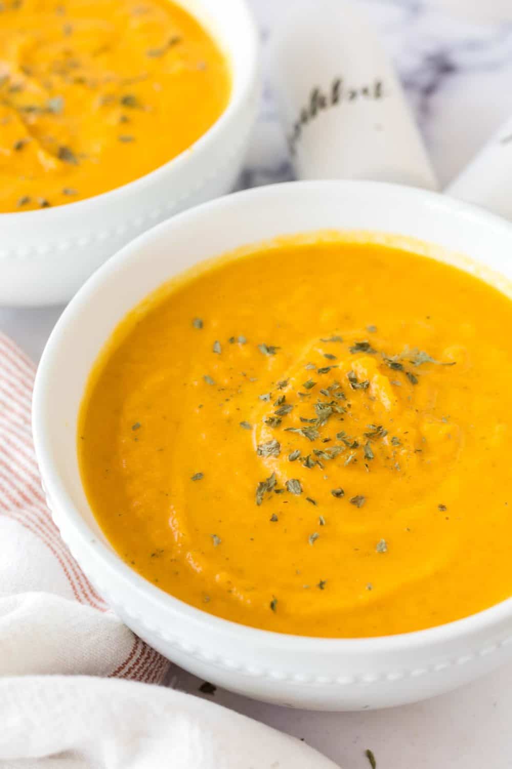 bright orange carrot soup in white serving bowls with herbs and salt and pepper to the side