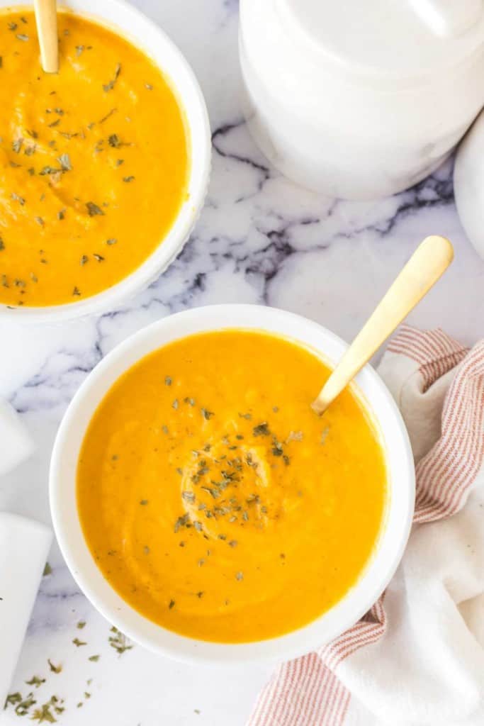 bright orange carrot soup in white serving bowls with herbs and salt and pepper to the side