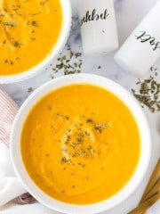 bright orange carrot soup in white serving bowls with herbs and salt and pepper to the side