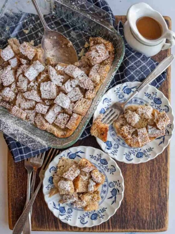 clear baking dish of squares of bread pudding with powdered sugar atop and served onto classic china dishes on the side
