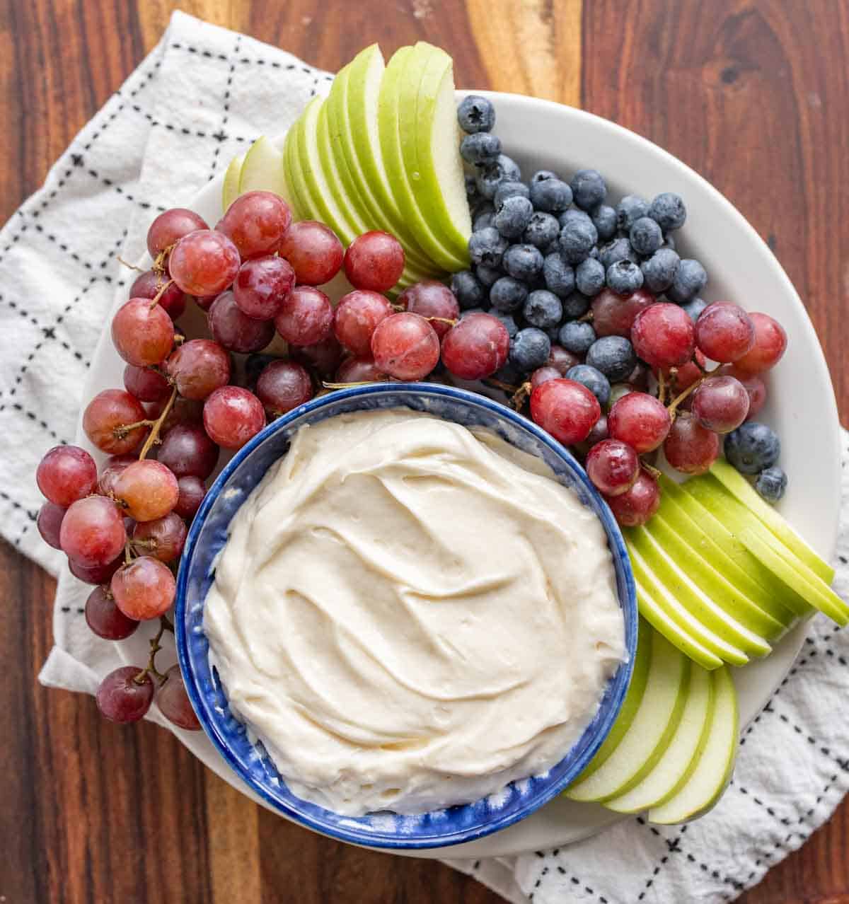 top view of fruit dip in a bowl with grapes blueberries and green apples sliced around it