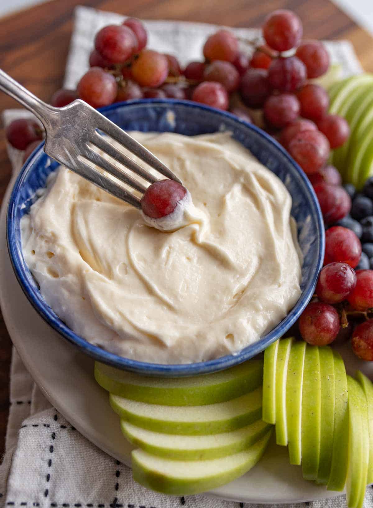 fruit dip in a bowl with grapes blueberries and green apples sliced around it with a fork dipping in a grab