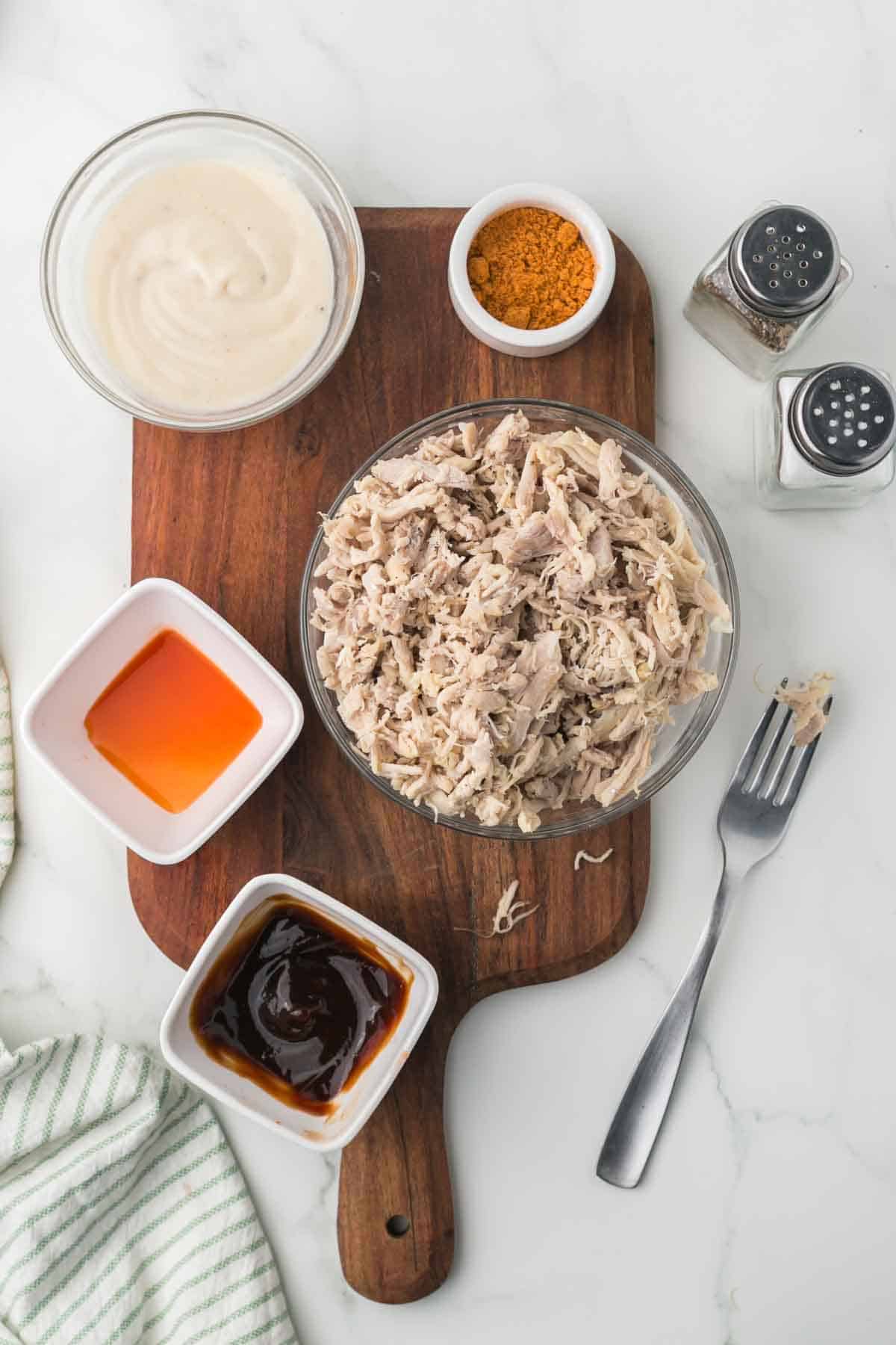 top view of shredded chicken in a clear bowl with seasonings and sauces around it
