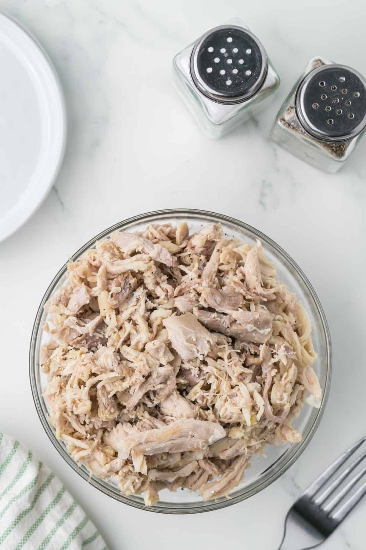 top view of shredded chicken in a clear bowl with seasonings and sauces around it