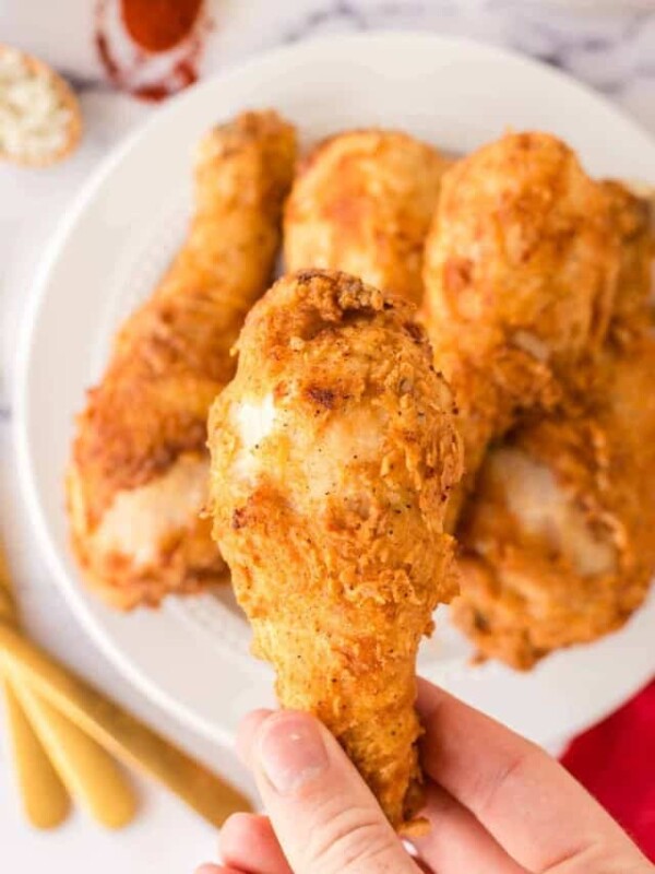 POV with a hand holding a leg of chicken over a white plate stacked with Louisiana chicken legs