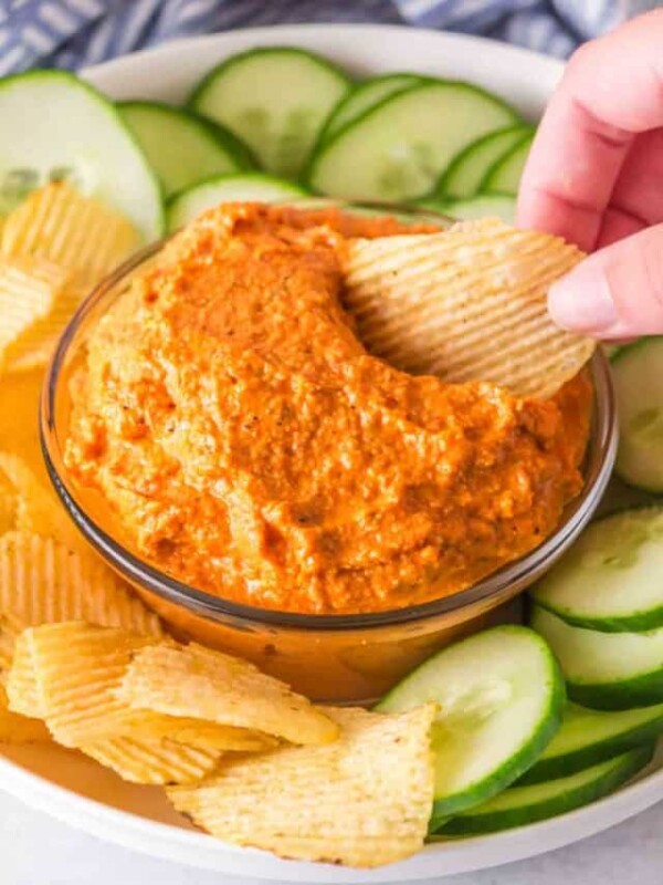 small dish with romesco sauce with crackers and chips and cucumbers around.