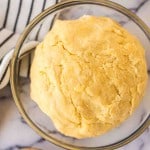 A ball of yellow empanada dough sits in a clear glass bowl on a marble surface, next to a black-and-white striped towel and a wooden spoon—perfect for your empanada dough recipe.