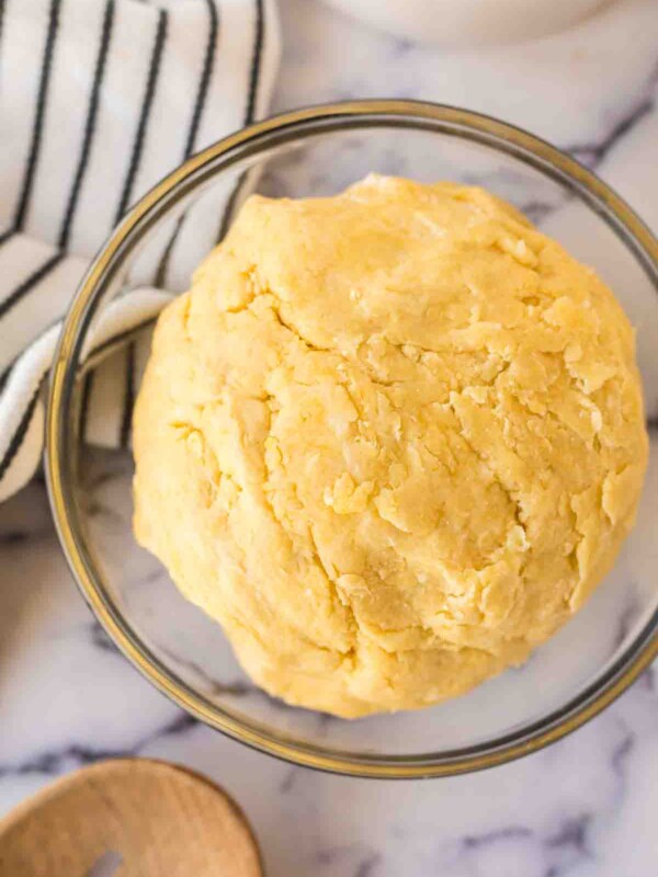 A ball of yellow empanada dough sits in a clear glass bowl on a marble surface, next to a black-and-white striped towel and a wooden spoon—perfect for your empanada dough recipe.