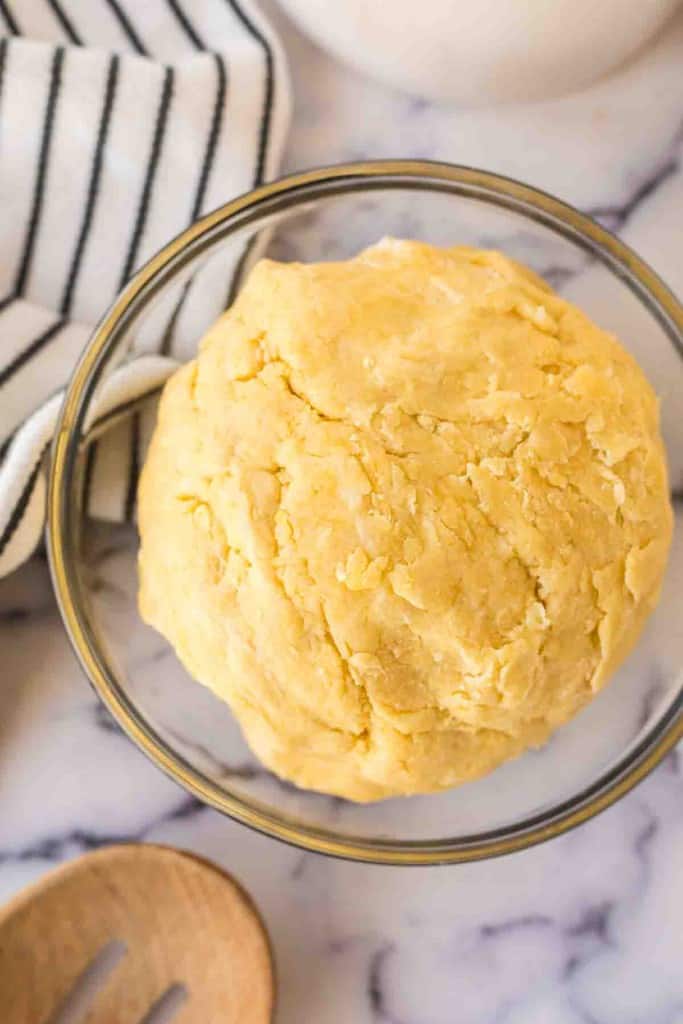 A ball of yellow empanada dough sits in a clear glass bowl on a marble surface, next to a black-and-white striped towel and a wooden spoon—perfect for your empanada dough recipe.