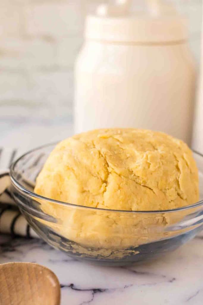 A ball of yellow empanada dough, made from a classic empanada dough recipe, sits in a clear glass bowl on a marble countertop, with a jar and a striped cloth in the background.