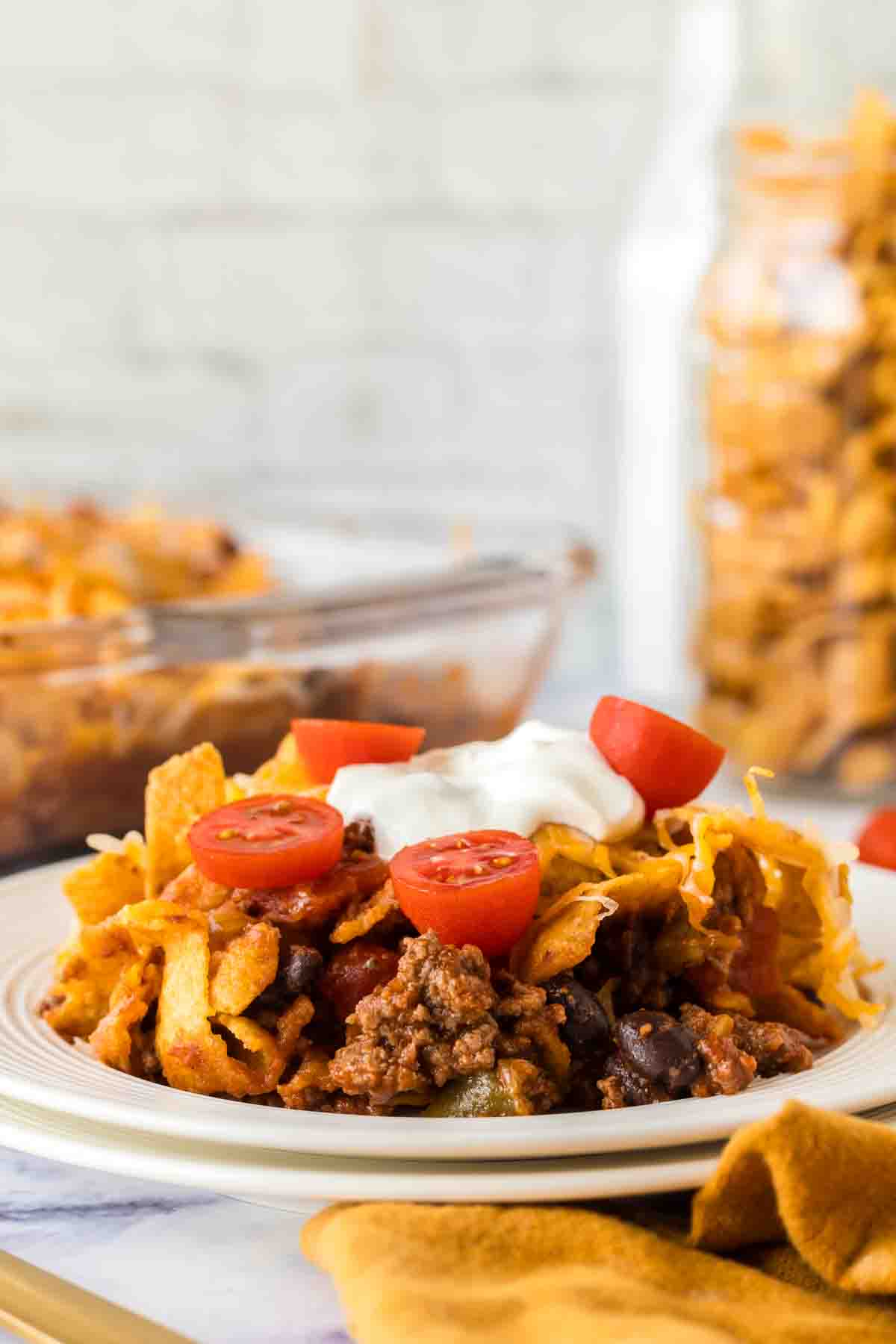 side view of a white dinner plate with frito pie with a dollop of sour cream and sliced cherry tomatoes on top