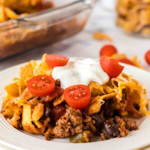 white dinner plate with frito pie with a dollop of sour cream and sliced cherry tomatoes on top