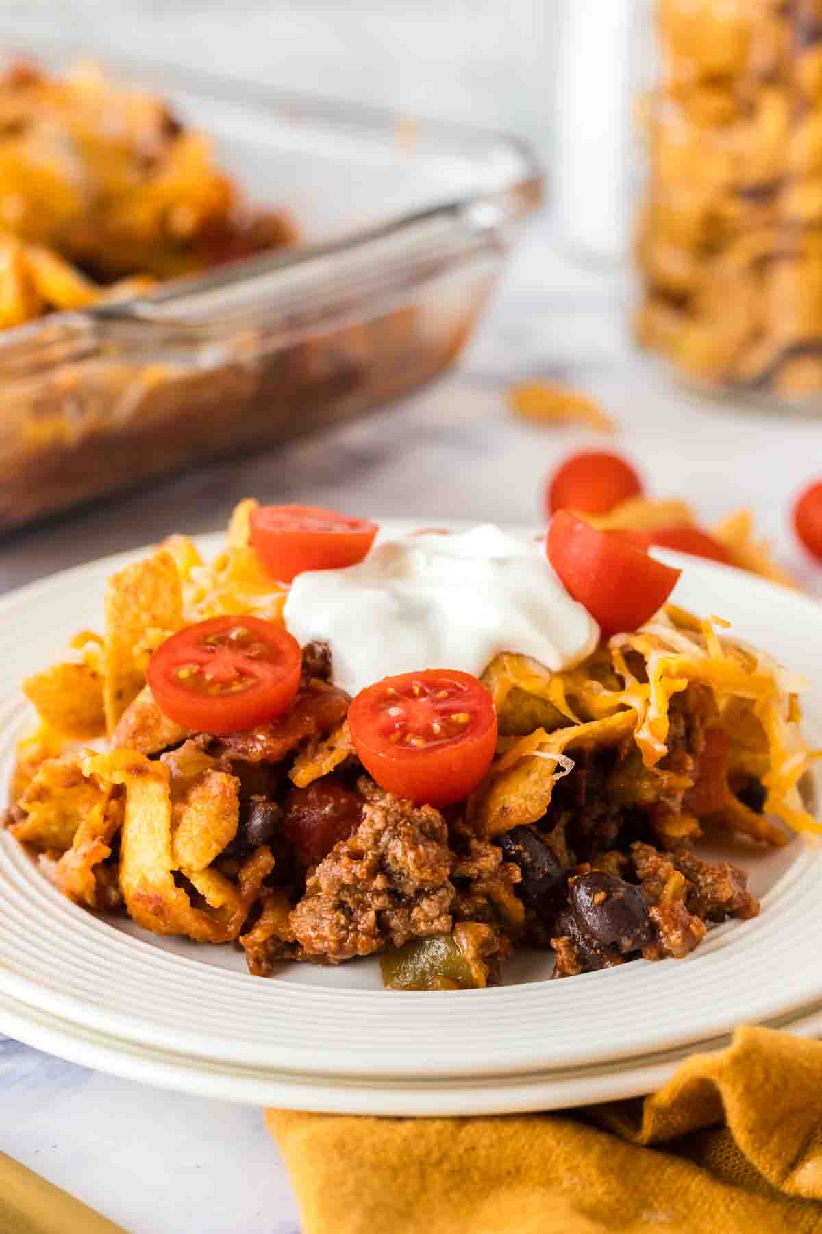white dinner plate with frito pie with a dollop of sour cream and sliced cherry tomatoes on top