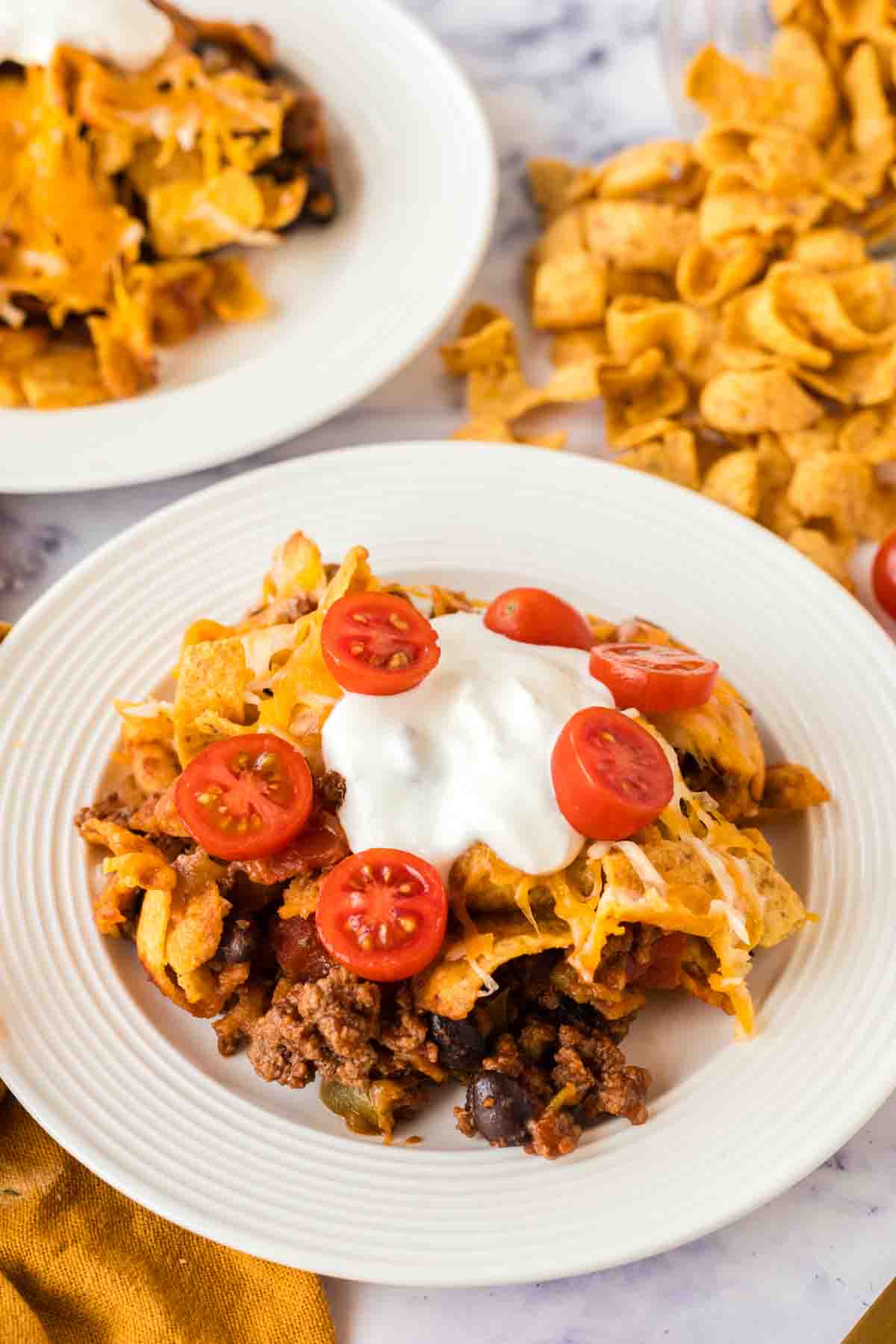 white dinner plate with frito pie with a dollop of sour cream and sliced cherry tomatoes on top