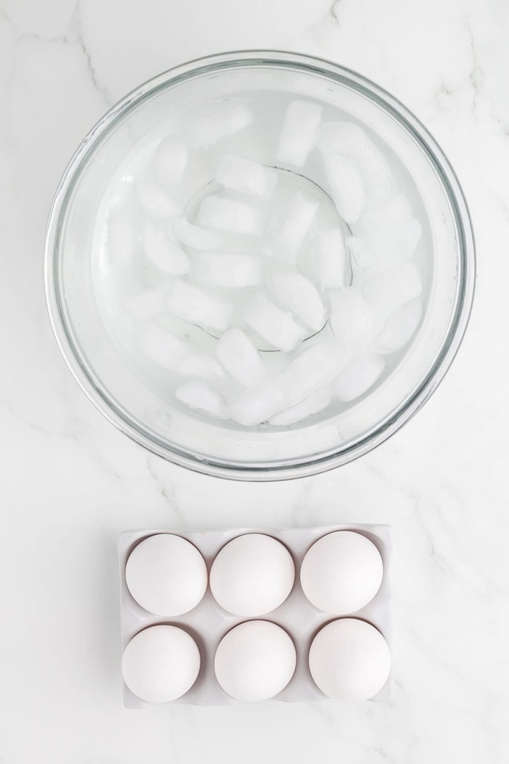 a clear mixing bowl of ice water next to a half dozen eggs laying on the table