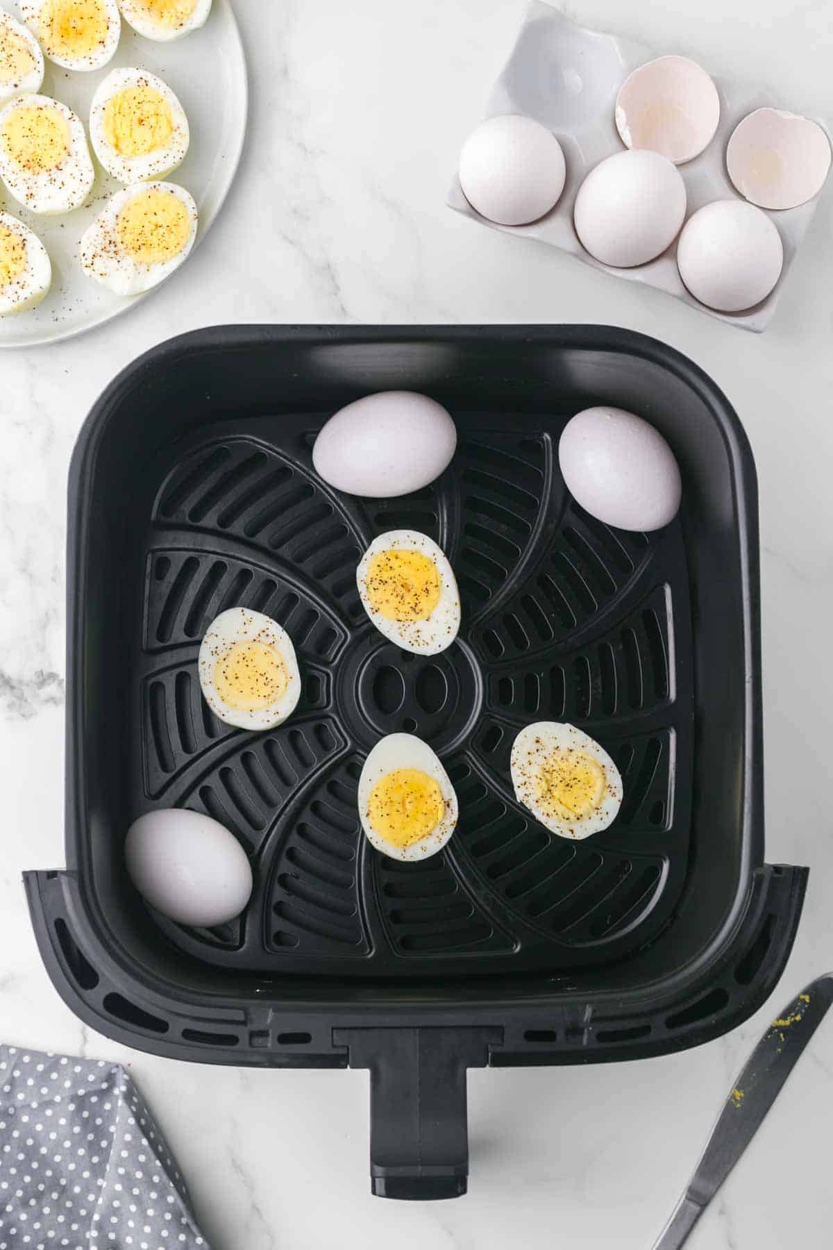 air fryer basket image with some whole eggs and some hard boiled eggs seasoned and cut in half