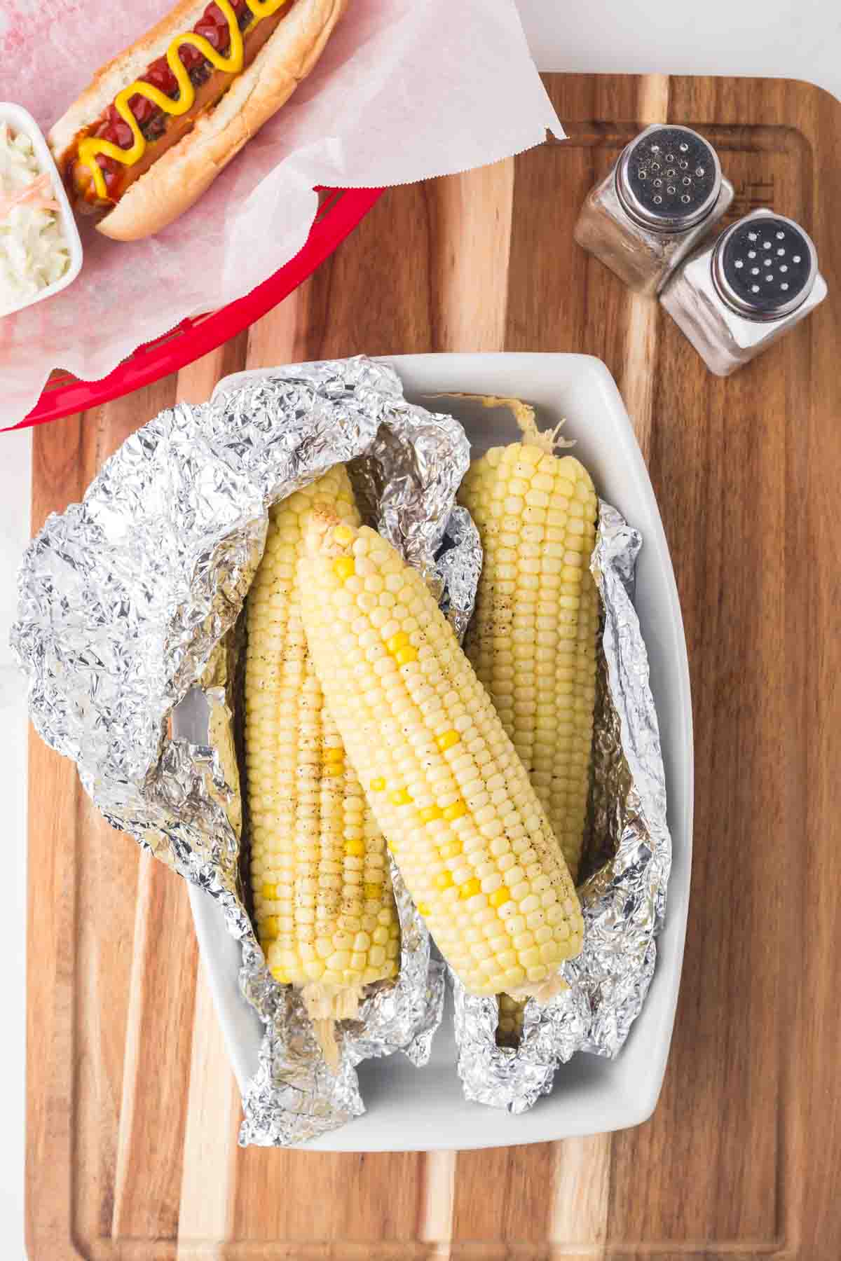 three pieces of corn on the cob wrapped in foil laying on a wooden board then the foil is opened