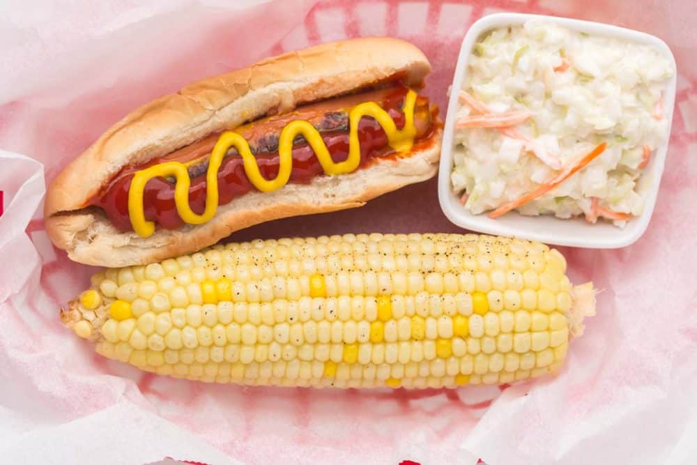 a paper lined basked with a classic hot dog with ketchup and mustard with a dish of coleslaw and a cooked corn on the cob