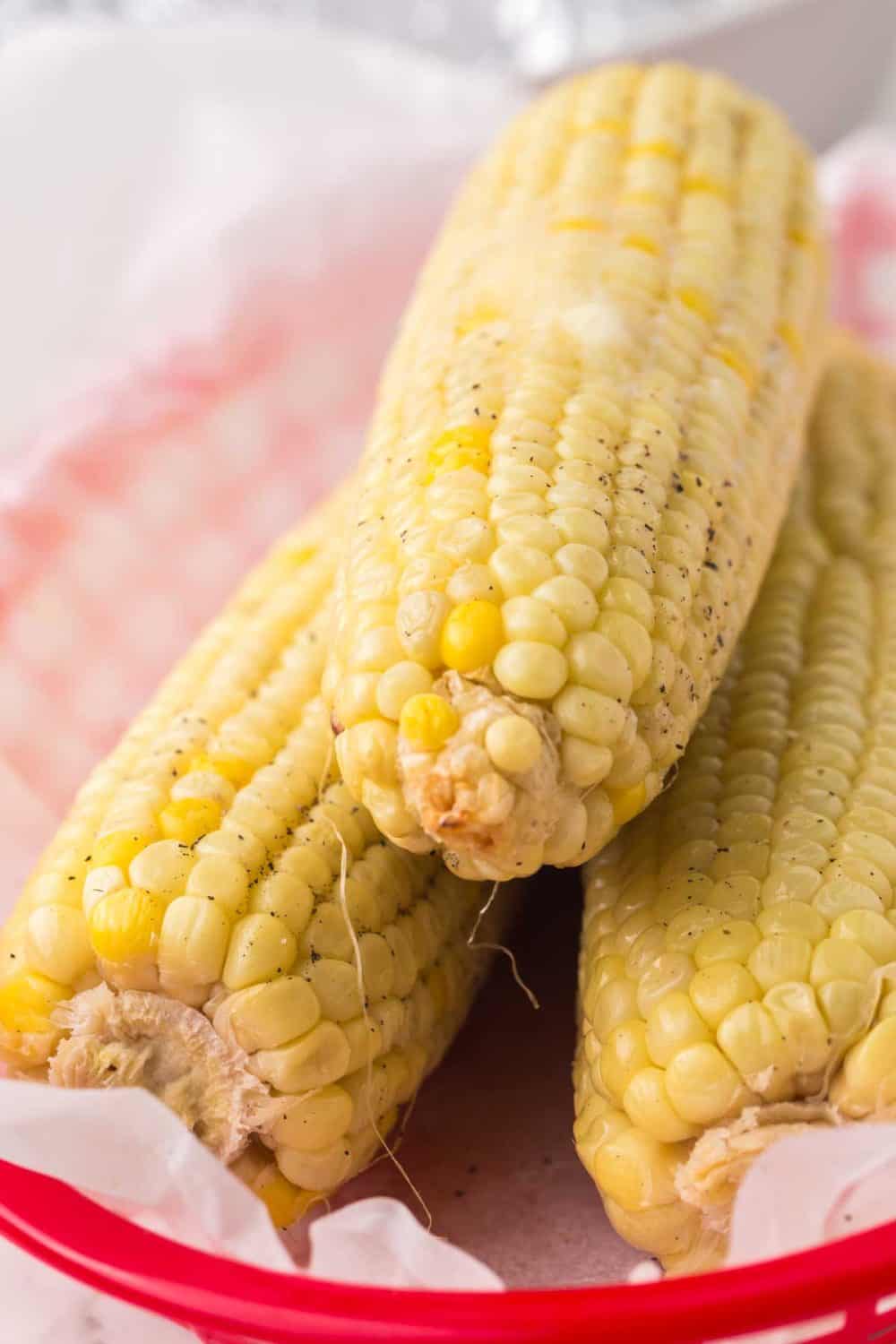 side view image of a paper lined basked with three cooked and buttered corn on the cobs stacked inside
