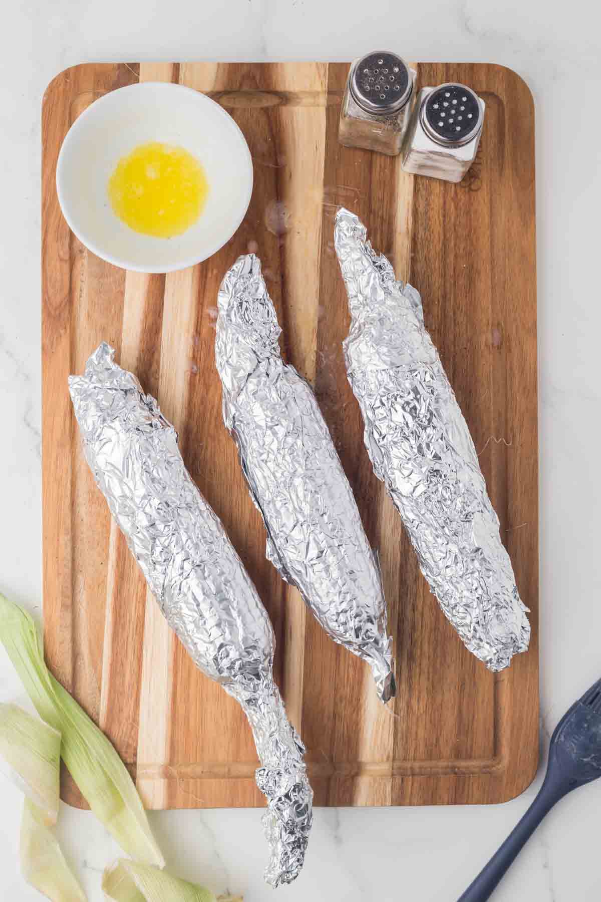 three pieces of corn on the cob wrapped in foil laying on a wooden board