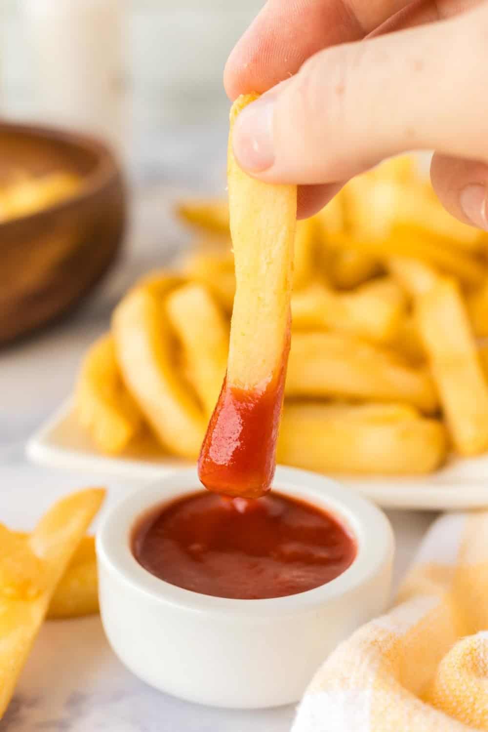 Hand dipping a fry into ketchup next to a long rectangle plate of air fryer french fries.