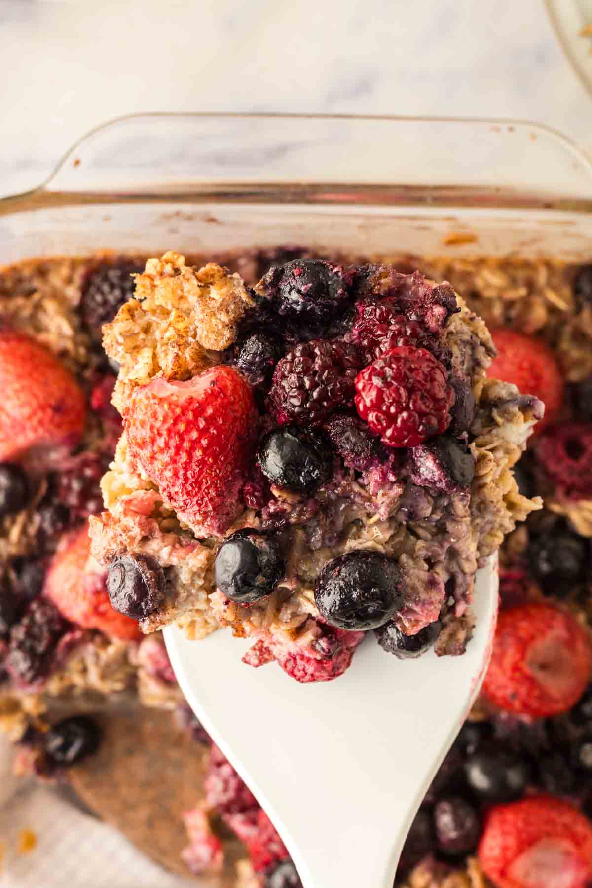 clear square casserole dish with baked oatmeal berries baked to a golden brown with a white spatula