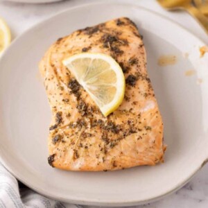 baked salmon filet on a round plate with a thin lemon slice on top.