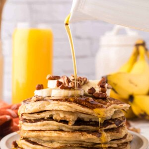 side view of a stack of banana pancakes on a round plate with walnuts and bananas with syrup being poured on top