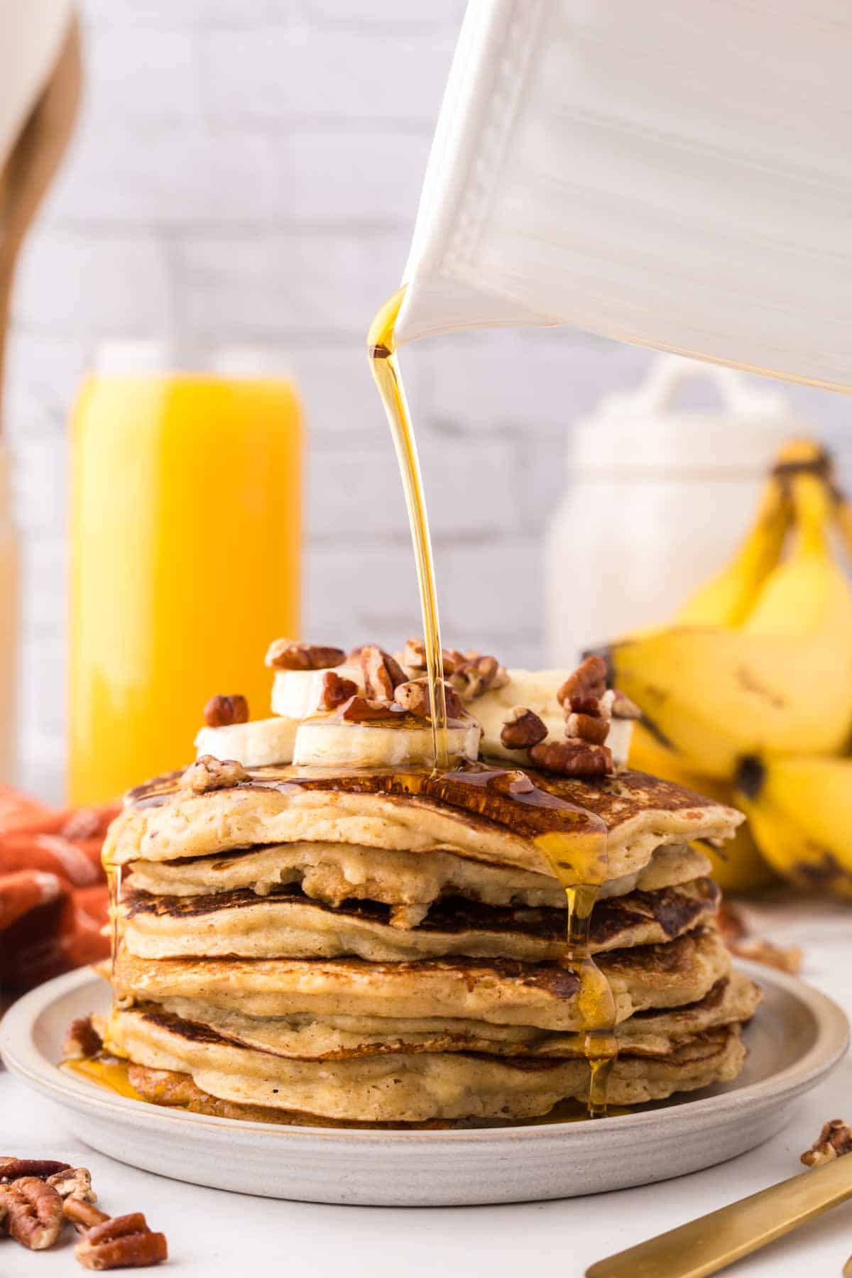 side view of a stack of banana pancakes on a round plate with walnuts and bananas with syrup being poured on top