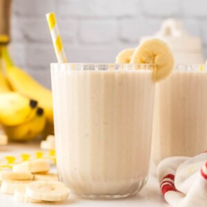 side view of a ribbed glass with a creamy banana smoothie with fresh bananas and a yellow striped paper straw two others in th backgrounders