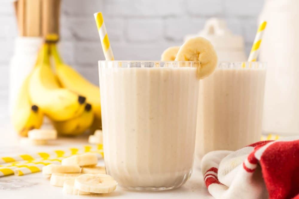 side view of a ribbed glass with a creamy banana smoothie with fresh bananas and a yellow striped paper straw two others in the background.