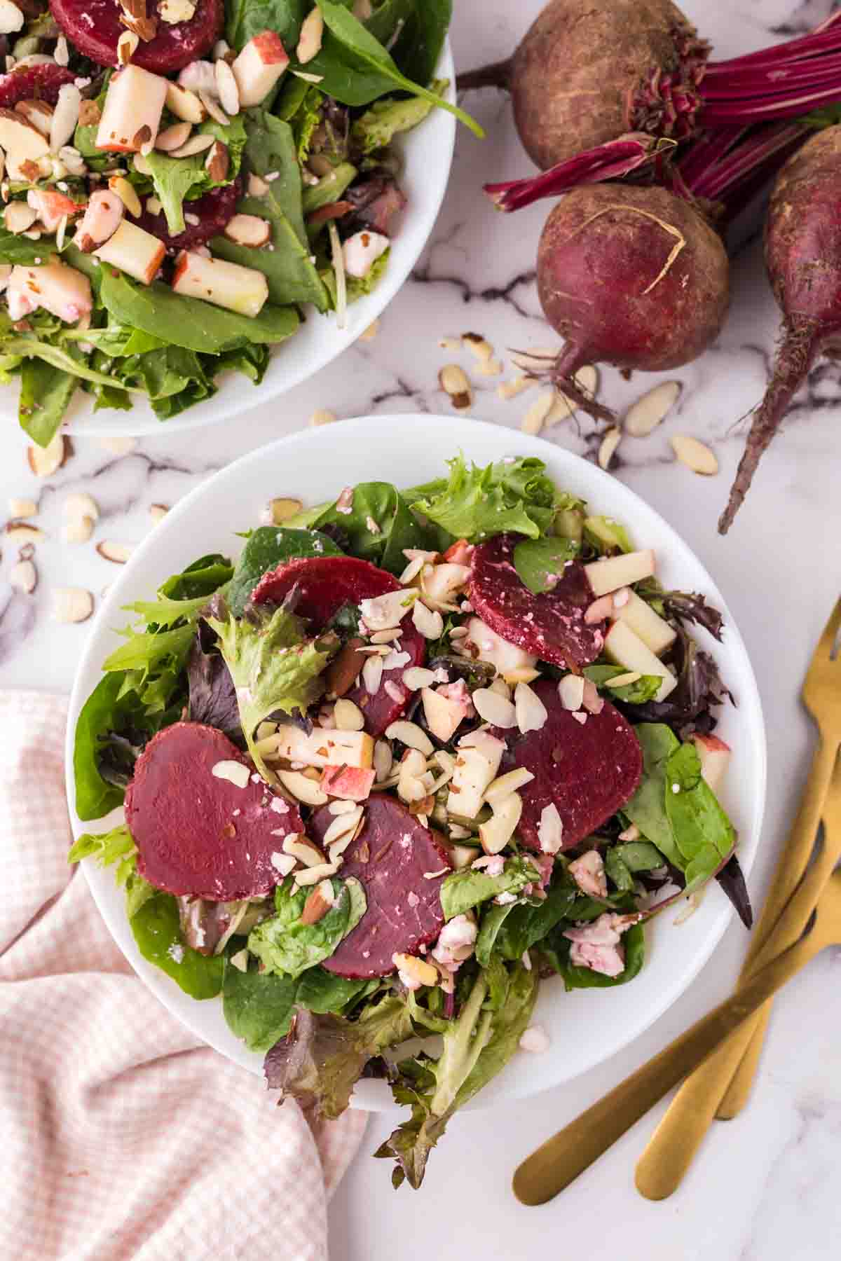 Beet salad with almonds and apples on top in a white bowl with golden forks to the side.